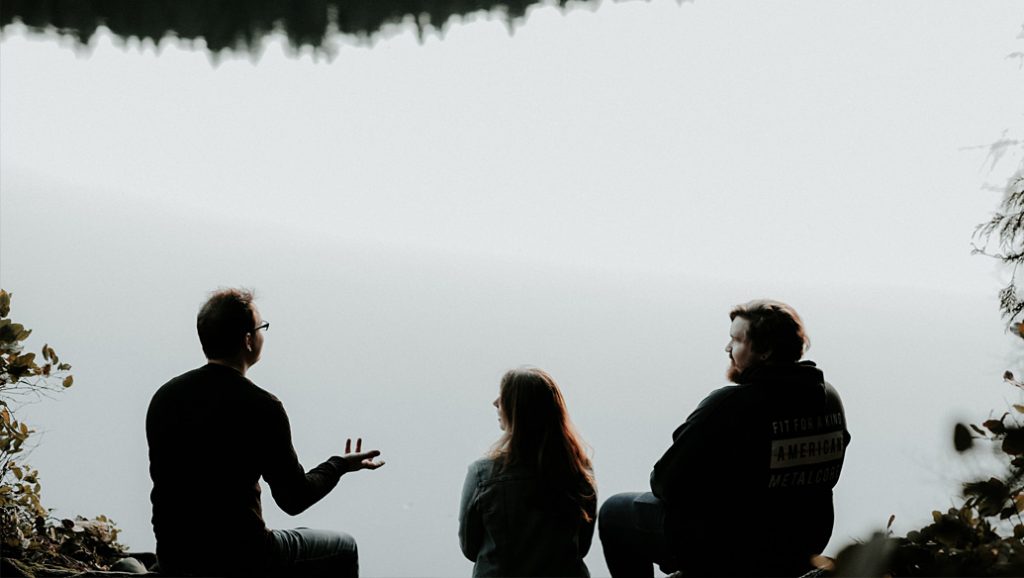 Two man and a woman talk while sitting beside a lake.