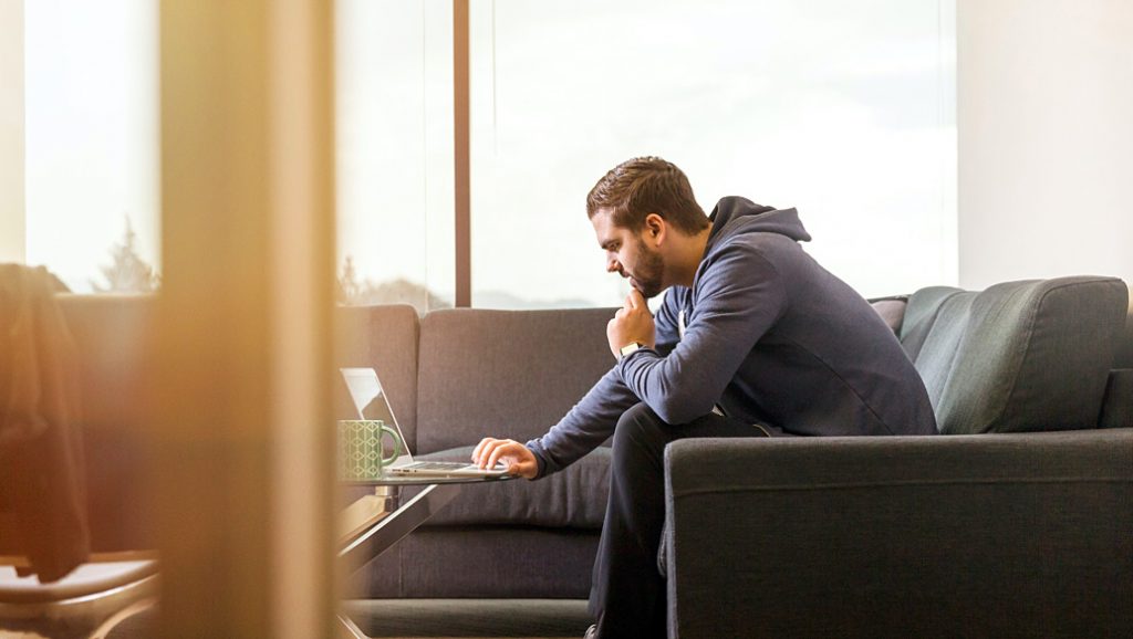 Man in hoodie sitting on a couch working on his laptop.