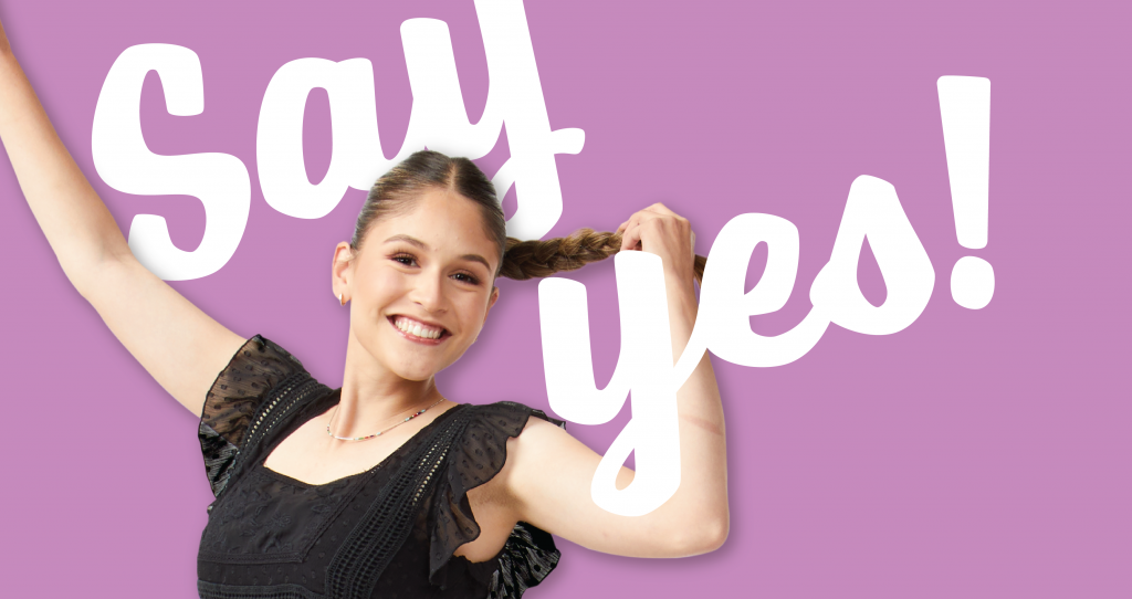A woman with a ponytail in a black dress raising her arms in front of a pink background with the words, "Say Yes!" overlaid.