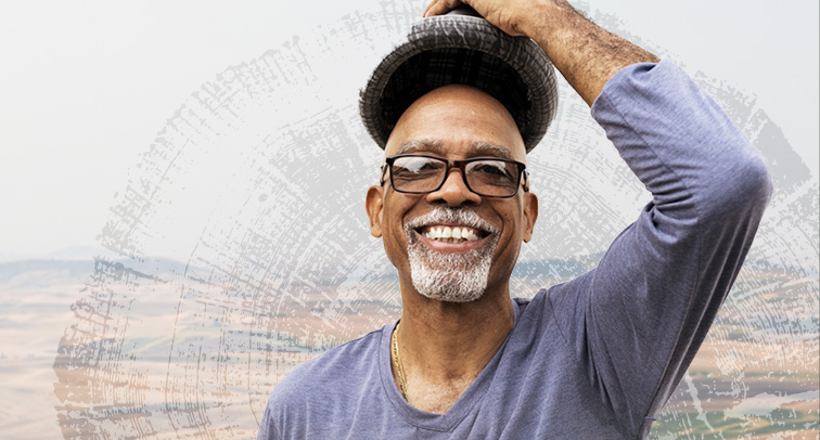 A smiling man in glasses lifts his hat off his head outside.